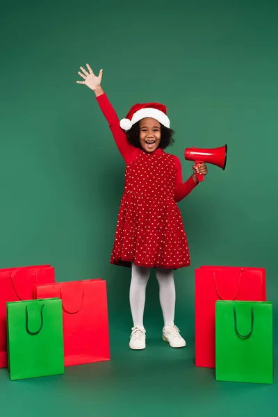 Positive african american girl in santa hat holding loudspeaker near shopping bags on green background — Stock Photo