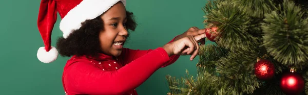 Positive afrikanisch-amerikanische Kind mit Weihnachtsmütze berührt Ball auf Weihnachtsbaum isoliert auf grün, Banner — Stockfoto