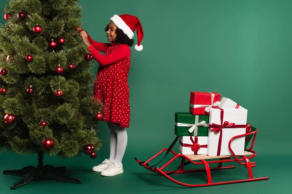 Side view of smiling african american girl in santa hat decorating christmas tree near sleigh with gifts on green background — Stock Photo