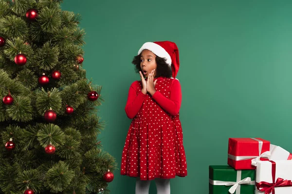 Niño afroamericano asombrado en sombrero de santa mirando el árbol de Navidad cerca de los regalos sobre fondo verde - foto de stock