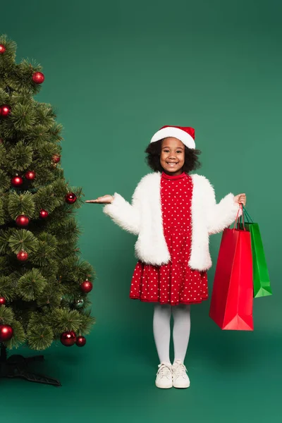 Happy african american girl in santa hat and fluffy jacket holding shopping bags and pointing at christmas tree on green background — Stock Photo