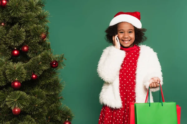Cheerful african american girl in santa hat holding shopping bags and talking on smartphone near christmas tree isolated on green — Stock Photo