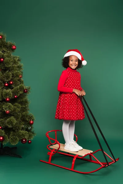 Sonriente niño afroamericano en vestido y sombrero de santa de pie en trineo cerca del árbol de Navidad sobre fondo verde - foto de stock