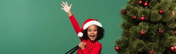 Positive afrikanisch-amerikanische Mädchen mit Weihnachtsmütze winkt in die Kamera in der Nähe von Weihnachtsbaum isoliert auf grün, Banner — Stockfoto