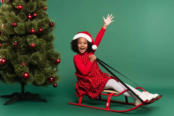 Emocionado afroamericano chica en santa hat saludando a cámara en trineo cerca de árbol de Navidad sobre fondo verde - foto de stock