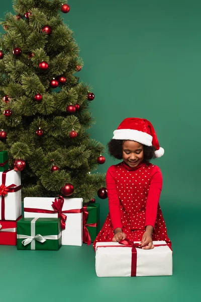 African american girl in santa hat holding bow on present near christmas tree on green background — Stock Photo