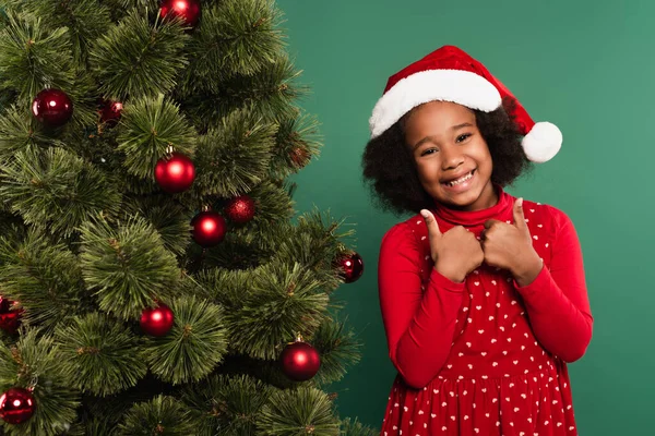 Niño afroamericano positivo en sombrero de santa rojo mostrando pulgares hacia arriba cerca del árbol de Navidad sobre fondo verde - foto de stock