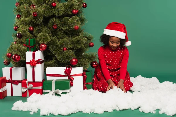 Afrikanisches Kind mit Weihnachtsmütze mit dekorativem Schnee in der Nähe von Weihnachtsbaum und Geschenken auf grünem Hintergrund — Stockfoto