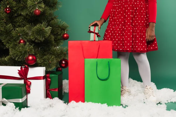 Vista cortada da criança americana africana colocando presente no saco de compras na neve decorativa perto da árvore de natal no fundo verde — Fotografia de Stock