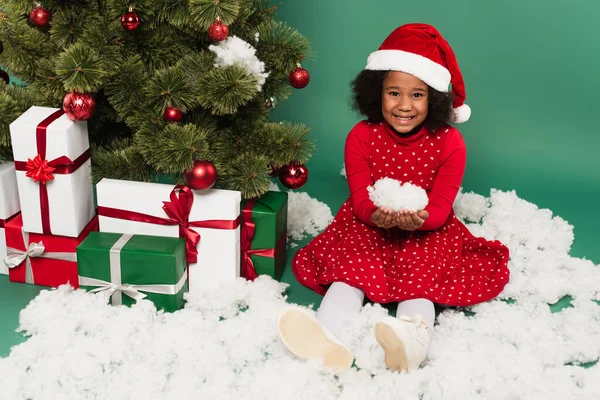 African american kid in santa hat holding decorative snow near presents and christmas tree on green background — Stock Photo