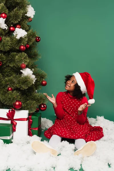 African american kid in santa hat pointing at christmas tree with gifts on decorative snow on green background — Stock Photo