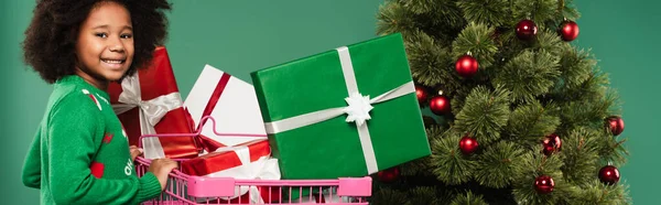 Smiling african american kid in sweater standing near shopping cart with gifts near christmas tree isolated on green, banner — Stock Photo