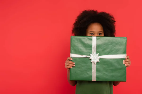 African american kid holding present near face isolated on red — Stock Photo