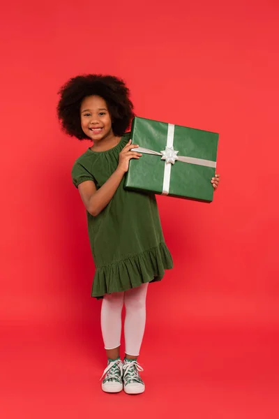 Glückliche afrikanisch-amerikanische Mädchen in Kleid mit Geschenk und Blick in die Kamera auf rotem Hintergrund — Stockfoto