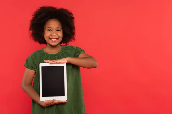 Positive afrikanisch-amerikanische Mädchen im Kleid hält digitale Tablette mit leerem Bildschirm isoliert auf rot — Stockfoto