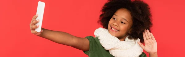 Smiling african american child in warm scarf having video call on smartphone isolated on red, banner — Stock Photo