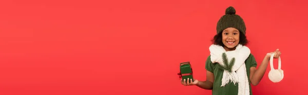 Menina afro-americana em chapéu e ramo de abeto em cachecol segurando caixa de presente e abafadores de orelha isolados em vermelho, banner — Fotografia de Stock