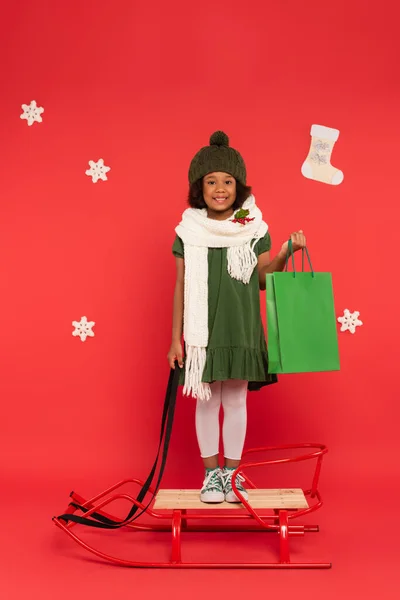 Chica afroamericana positiva en sombrero y bufanda con copos de nieve sosteniendo bolsa de compras en trineo cerca de copos de nieve sobre fondo rojo - foto de stock