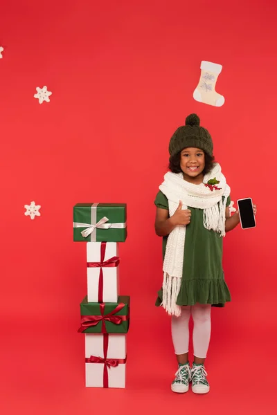 Positivo bambino afroamericano che tiene lo smartphone e mostra come gesto vicino a regali e fiocchi di neve su sfondo rosso — Foto stock