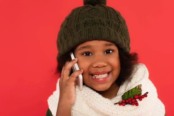 Happy african american girl in hat and scarf with mistletoe talking on mobile phone isolated on red — Stock Photo