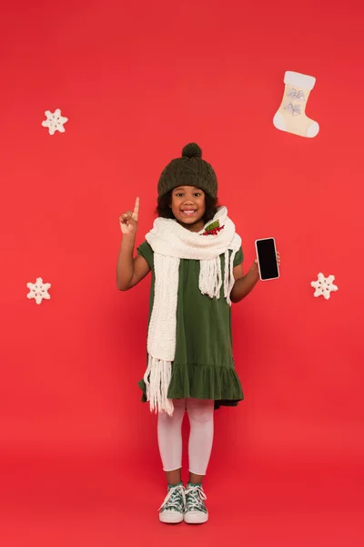 Sonriente niño afroamericano en bufanda con muérdago sosteniendo teléfono inteligente y teniendo idea cerca de copos de nieve sobre fondo rojo - foto de stock