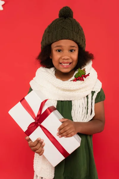 Cheerful african american kid with mistletoe on scarf holding gift box on red background — Stock Photo