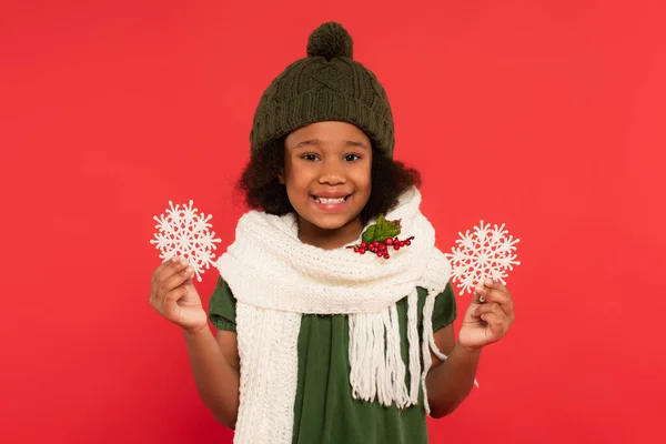 Sonriente chica afroamericana en sombrero y bufanda con muérdago sosteniendo copos de nieve decorativos aislados en rojo - foto de stock