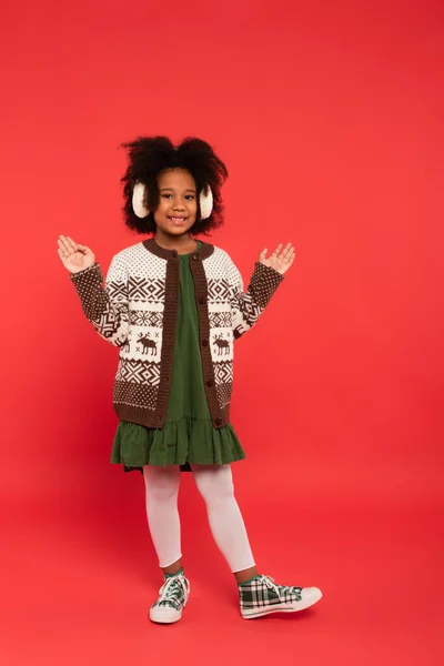 Enfant afro-américain dans des cache-oreilles et un cardigan pointant avec les mains sur fond rouge — Photo de stock