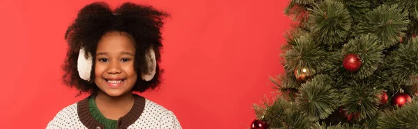 Afro-americana chica en orejeras sonriendo a cámara cerca de árbol de Navidad aislado en rojo, pancarta - foto de stock