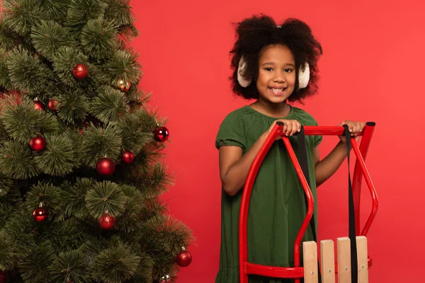 Smiling african american kid in ear muffs standing near sleigh and christmas tree isolated on red — Stock Photo