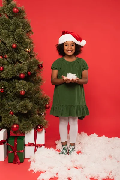 African american child in santa hat holding decorative snow near christmas tree and presents on red background — Stock Photo