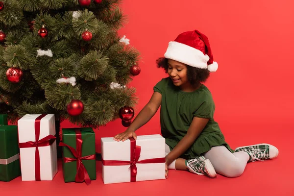 Happy african american girl in santa hat holding present near decorated christmas tree on red background — Stock Photo