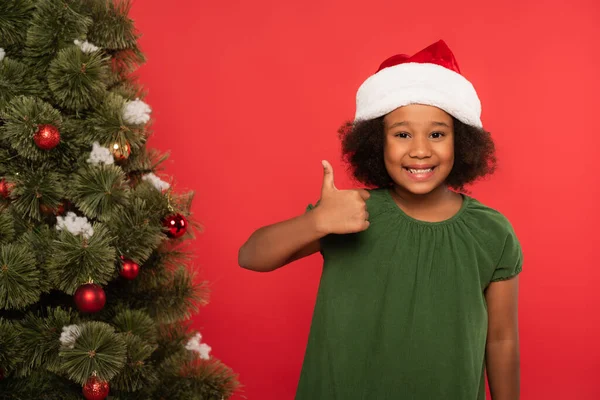 Positivo afroamericano ragazza in cappello di Babbo Natale mostrando come vicino albero di Natale isolato sul rosso — Foto stock