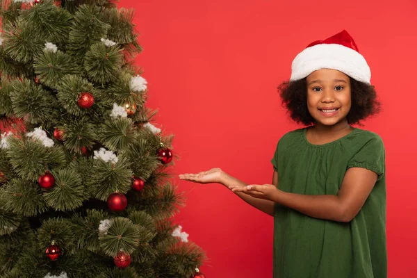 Lächelndes afrikanisch-amerikanisches Kind mit Weihnachtsmütze zeigt auf Weihnachtsbaum auf rotem Grund — Stockfoto