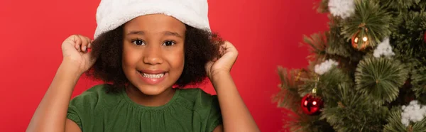 Bambino afroamericano con cappello vicino all'albero di Natale sfocato isolato sul rosso, striscione — Foto stock