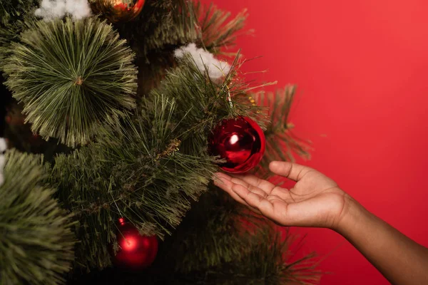 Vista recortada del niño afroamericano sosteniendo la pelota en el árbol de Navidad aislado en rojo - foto de stock