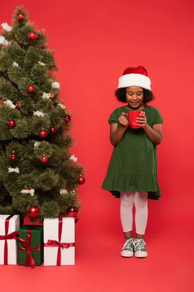 Afroamerikanerin mit Weihnachtsmütze hält Tasse in der Nähe von Weihnachtsbaum und Geschenkschachteln auf rotem Hintergrund — Stockfoto