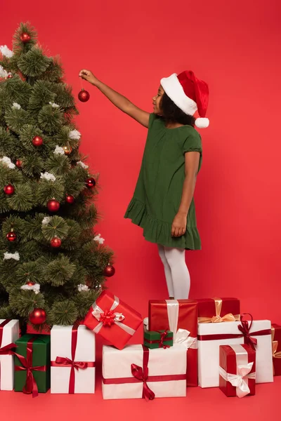 Vista lateral del niño afroamericano en santa hat sosteniendo pelota cerca del árbol de navidad y regalos sobre fondo rojo - foto de stock