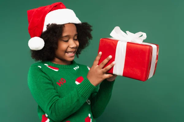 Positive african american girl in santa hat and sweater holding present isolated on green — Stock Photo