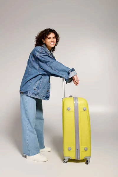 Charming woman in denim outfit stands beside vibrant yellow bag in a stylish background. — Photo de stock