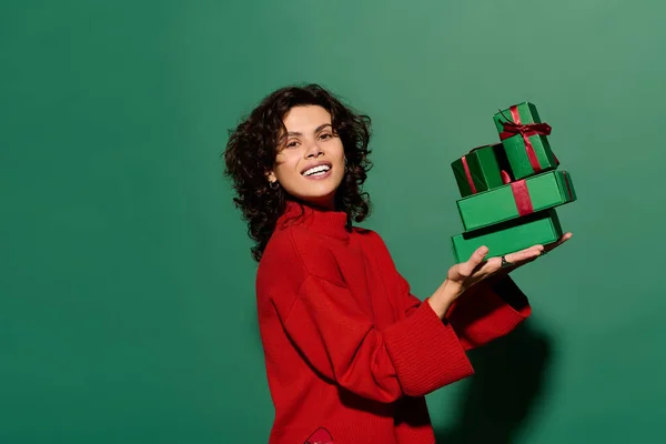 A woman in a red sweater smiles with wrapped gifts against a green backdrop. — стокове фото