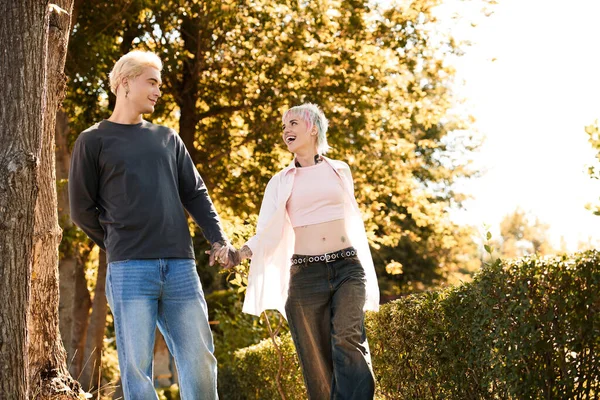 Una joven pareja camina de la mano a través de un parque iluminado por el sol, sonriendo alegremente el uno al otro. - foto de stock