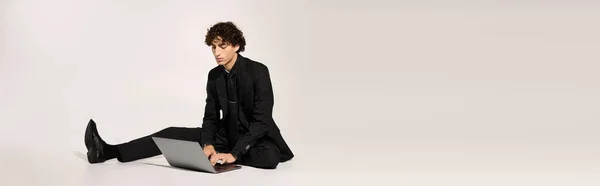 Стильний молодий чоловік сидить на підлозі, орієнтований на ноутбук, виключаючи впевненість і сучасну моду. — стокове фото