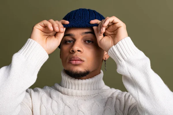 Schöner junger Mann im kuscheligen Pullover wechselt seine Mütze, während er für einen stylischen Look posiert. — Stockfoto