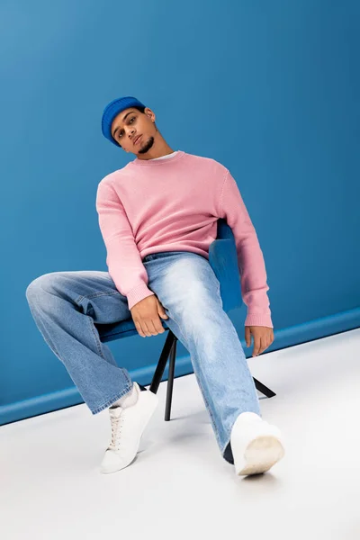 Стильный молодой человек в розовом свитере и джинсах небрежно позирует в ярко-синей обстановке. — стоковое фото