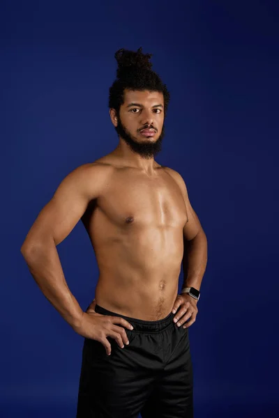 Подтянутый черный мужчина с вьющимися волосами и бородой уверенно позирует без рубашки на синем фоне. — стоковое фото