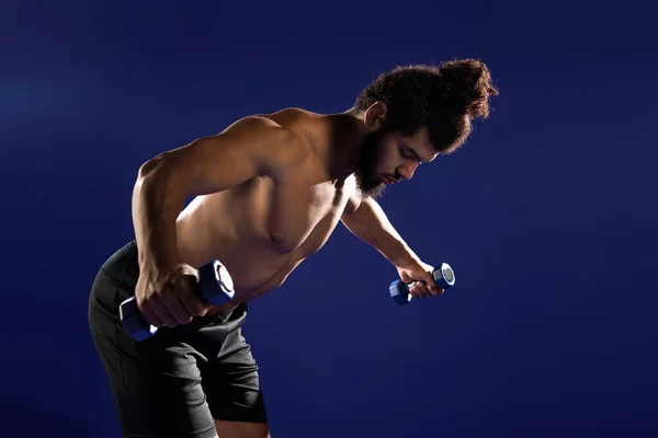 An athletic man focuses on his workout, performing exercises with dumbbells on a blue background. — Stock Photo