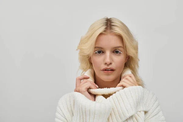 A beautiful young blonde woman wearing a cozy white sweater poses gracefully in a bright studio. — Stock Photo