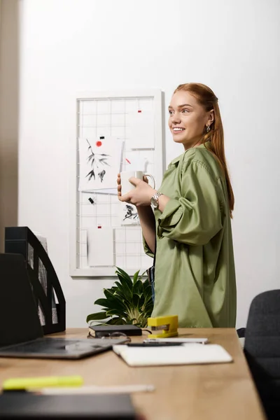 A young woman smiles warmly while holding a cup, surrounded by her inviting home workspace. — Stock Photo