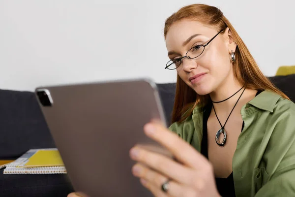 A young woman sits comfortably in her home, engrossed in her tablet while wearing stylish glasses. — Stock Photo
